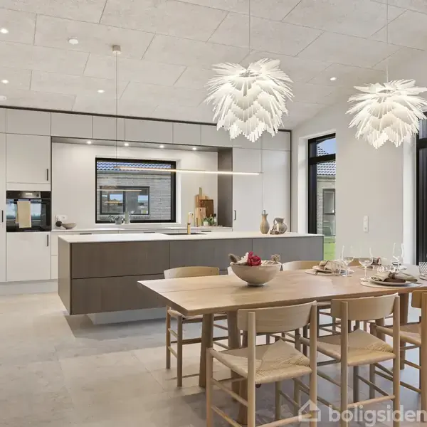Moderne køkken-alrum med et stort spisebord med træstole, placeret under to dekorative lamper. Køkkenet har hvidt skabsmøblement og central ø, set ud til en have gennem store vinduer.