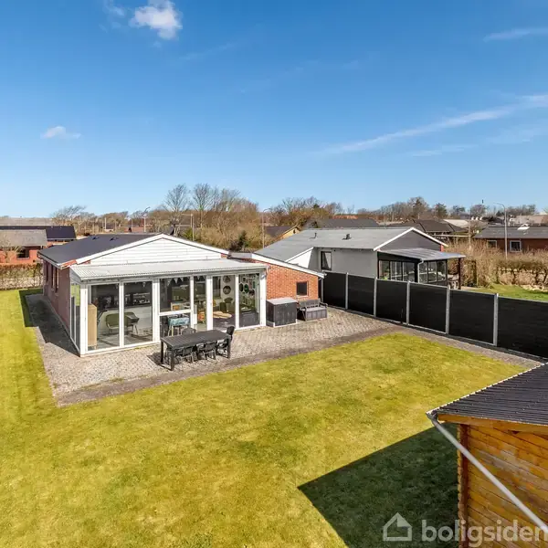 Et enkelt hus står i en velplejet have med tætklippede græsplæner og terrasse. Indrammet af hække og et sort hegn, omgivet af andre boliger i en rolig forstad.