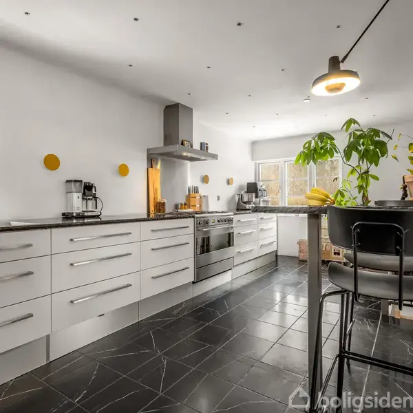 Køkken med hvide skabe og stålgreb, udfører funktion som madlavningsområde. Sort flisegulv og lys vægfarve skaber moderne atmosfære. Både planter og stole tilføjer hygge ved rund bordplade.