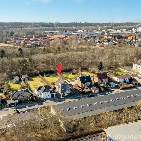 Hus fremhævet med rød pil, omgivet af andre beboelsesejendomme og træer i en landsby. Foran er der flade tage på industrielle bygninger. I baggrunden ses flere huse og skov.
