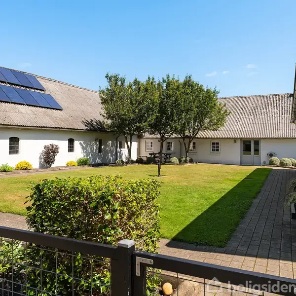 Hvid bygning med solpaneler på taget omgiver en grøn græsplæne med et træ i midten. Flisebelagt sti løber langs facaden. Der er klare blå himmel og dagslys.