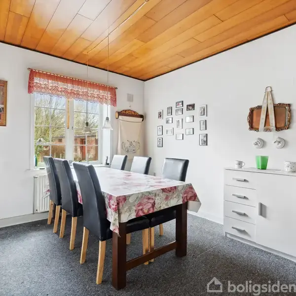 Langt spisebord med otte sorte stole omkring står i en lys spisestue. Væggene er dekoreret med billeder, og der er en kommode i højre side.