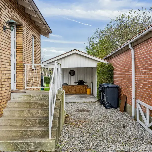 Trappeopgang fører op til en murstensbygning. To affaldscontainere står i grusområdet mellem husene. I baggrunden ses et skur med et bord og en vægmonteret ur.