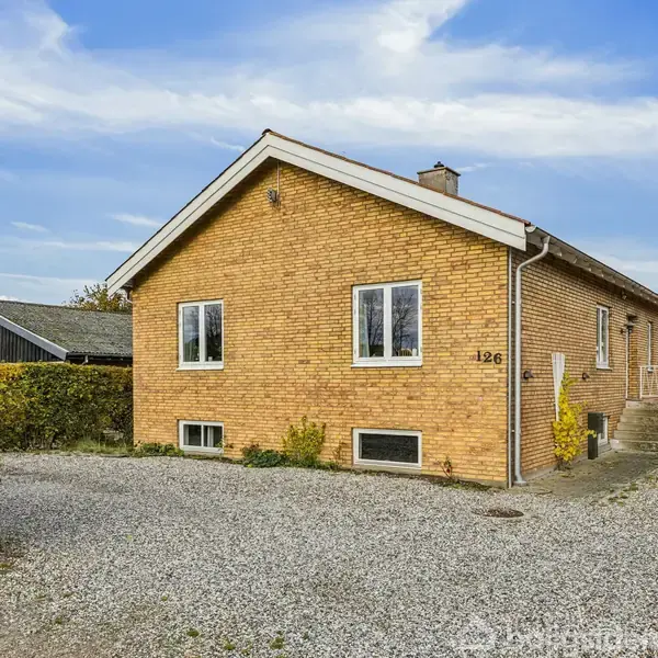Et gult murstenshus står stille i en grusbelagt indkørsel, omgivet af hæk. Husnummeret "126" ses tydeligt. I baggrunden er der en garage og en blå himmel med skyer.