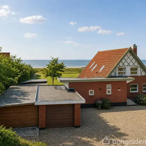Et rødt hus med en vinkelformet tagkonstruktion står stille foran en garage. Omgivet af træer med havudsigt i baggrunden og en grusbelagt indkørsel i forgrunden.