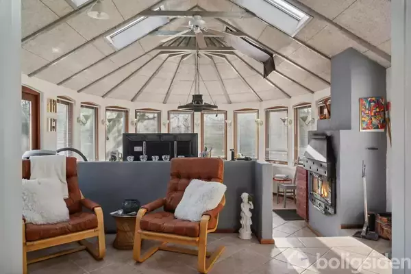 To lænestole med puder står i en stue med en buet glasvæg, hvor der er lysindfald fra ovenlysvinduer. En brændeovn er indbygget i væggen.