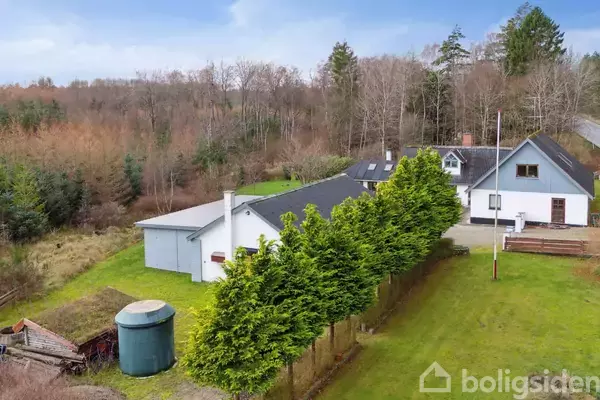 Et parcelhus med en stor grøn gårdsplads omgivet af høje træer og buske i et skovområde. Til højre er der et hvidt hus og en garagebygning.