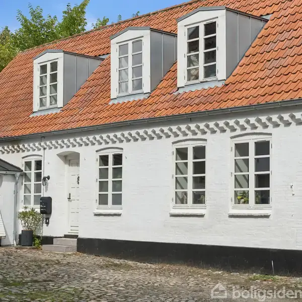 Et hvidt murstenshus med rødt tegltag har tre loftsvinduer og klassiske døre mod en brostensbelagt gårdsplads omgivet af grønne træer.