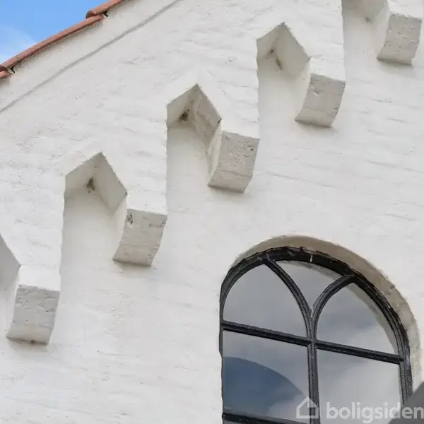 Hvid bygning med takkede murstensdetaljer og en buet, sortrammet vindue. Kontekst er en klar himmel med få skyer. Vandmærke "boligsiden" nederst til højre.