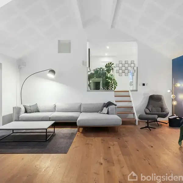 En moderne stue med en grå sofa og et sofabord, beliggende i et rum med trægulv. En buet gulvlampe står ved sofaen, og der er planter og en lænestol i baggrunden.