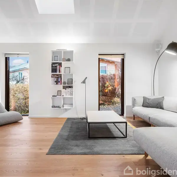 Stue med en grå sofa og gulvlampe. Sofabord på tæppe, hylde med bøger ved væggen. To døre viser udsigt til have. Lyse træplanker dækker gulvet.