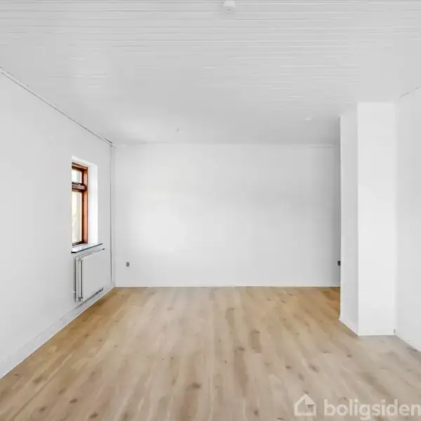 Et rektangulært, tomt rum med lyse trægulve, hvide vægge og et vindue til venstre. En radiator er placeret under vinduet. Vandmærket "boligsiden" er i nederste højre hjørne.