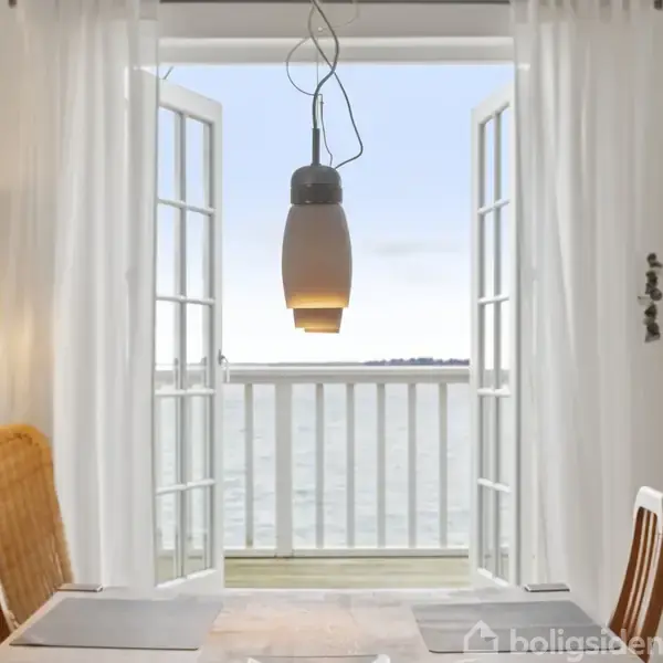 En spisebordslampe hænger over et bord, med stole omkring, foran åbne franske døre der fører ud til en balkon med udsigt over vandet. En sofa og væglampe står til venstre.