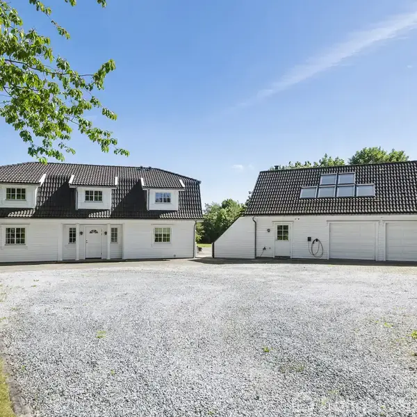 To hus med sort tag står på grusplads omgivet af grønne træer. Et hus har vinduer på taget og garage; det andet har flere kviste. Klar blå himmel ses.