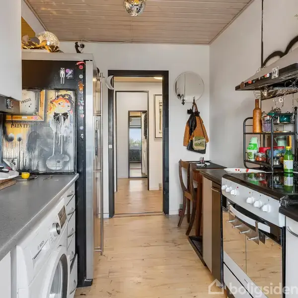 Køkken med hvide skabe og sort bordplade. En ovn og køleskab står til venstre, omgivet af husholdningsartikler. Bagved er en gang, der fører til et værelse.