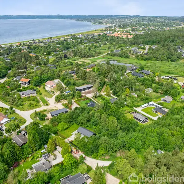 Luftfoto af et grønt boligområde med spredte huse omgivet af træer og haver. I baggrunden ses en kystlinje med roligt vand.