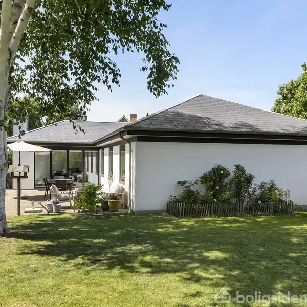Huset er en moderne hvid bungalow med sort tag. Foran huset er en velplejet græsplæne og et træ i venstre side. Haven er omkranset af hække.