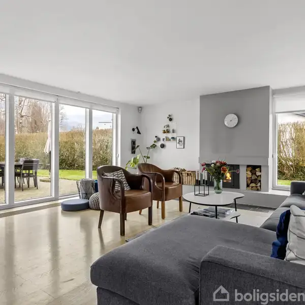 Stue med grå sofa og lænestole omkring et sofabord, pejs til højre med dekorative elementer. Stor glasskydedør leder til terrasse med havemøbler. Lyse vægge og trægulv.