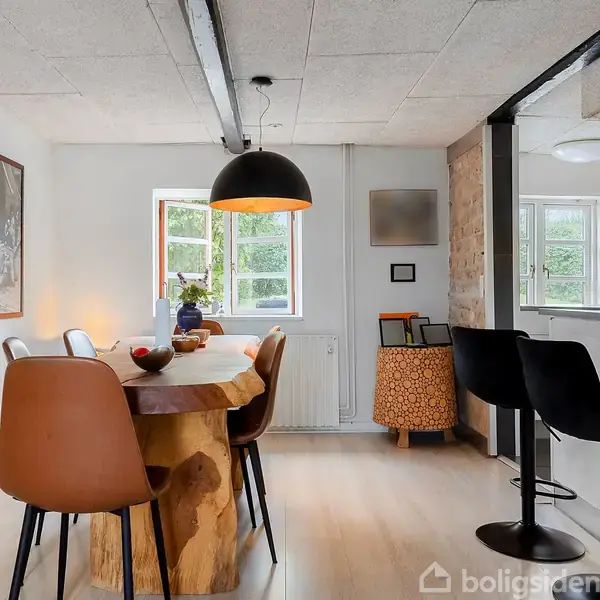 Et rundt spisebord af træ er omgivet af forskellige stole. Bordet står i et lyst rum med store vinduer, en stor lampe hænger over det, og kunst pryder væggen.