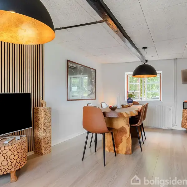 TV står på mønstret bord, slukket, i moderne stue med trædetaljer og spisebord. Stole omringer bordet, og store loftslamper hænger over det. Vindue bagved giver naturligt lys.
