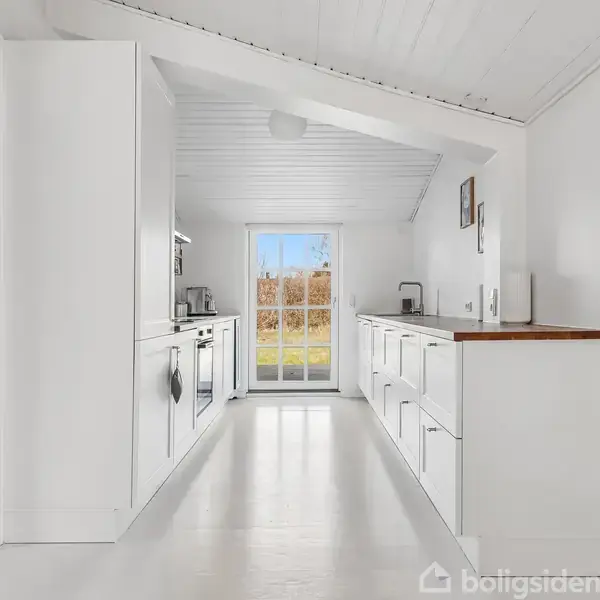 Et sort træhus står stille på en græsplæne omgivet af træer. Huset har hvide vinduer og en terrasse i træ foran. Himlen er blå med få skyer.