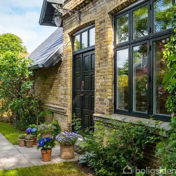 En murstensbygning med sort dør og vinduer står ved siden af en sti. Omkring er blomster i krukker og grønne planter, omkranset af en velplejet have med hæk og græs.