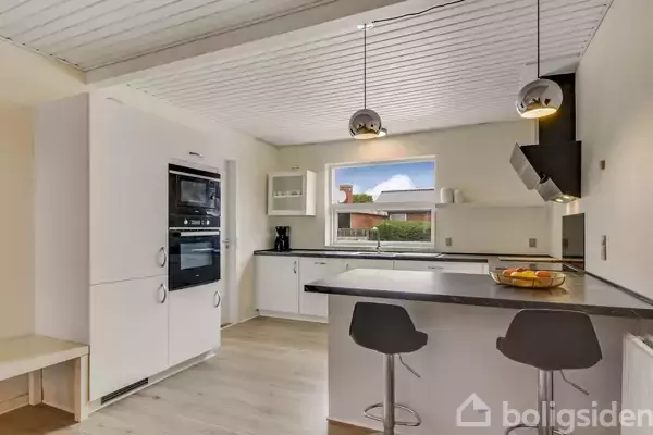 Et moderne køkken har hvide skabe, sorte barstole ved en sort køkkenø og to hængende lamper. Et stort vindue slipper dagslys ind, og frugtskål står på bordet.