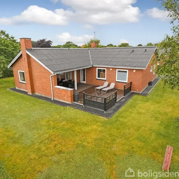 En rød murstensvilla med sort tag står i en velholdt, grøn have. Villaen har en træterrasse med liggestole og rækværk. Skyfri himmel og træer omkranser huset.