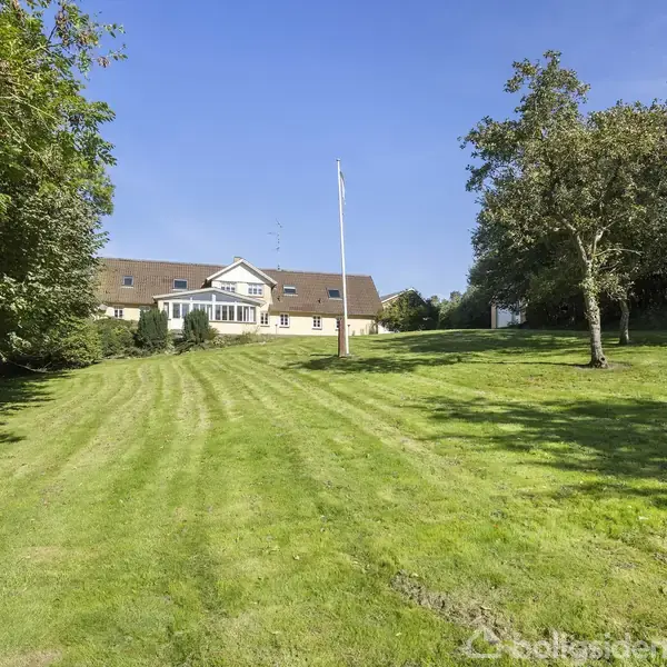 Et hvidt hus står i baggrunden med en stor, velplejet græsplæne foran. På plænen er der en flagstang og flere træer, omgivet af skov på begge sider.