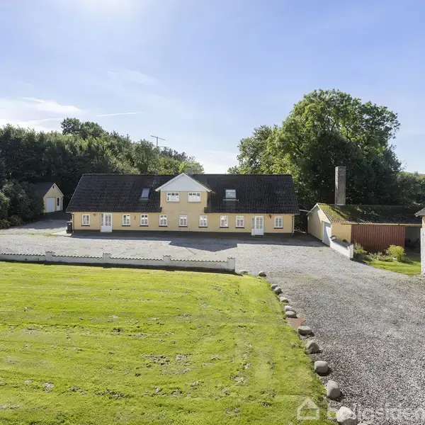 En gul landgård med mørkt tag ligger i en gårdsplads omringet af grusarealer og græs. Baglandet består af grønne træer under en klar blå himmel.
