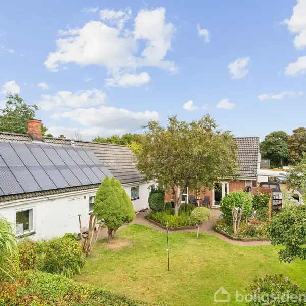 Et hvidt hus med solpaneler på taget står i en grøn have med træer og buske. Himlen er blå med spredte skyer.
