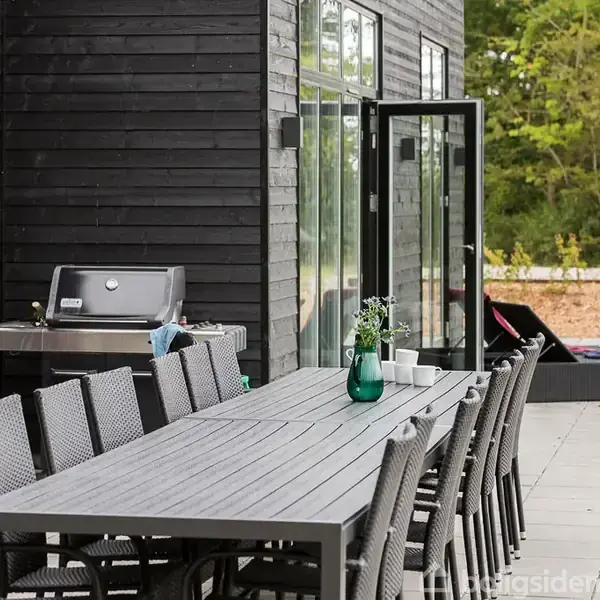 Langt havebord med ti stole står på en flisebelagt terrasse ved et sort træhus. Grillen står mod væggen. Omgivet af grøn natur.