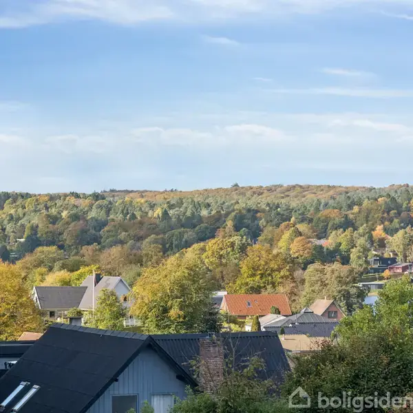 Huse ligger i en skovklædt dal, farvet af efterårets nuancer; skyer og blå himmel over trætoppene.