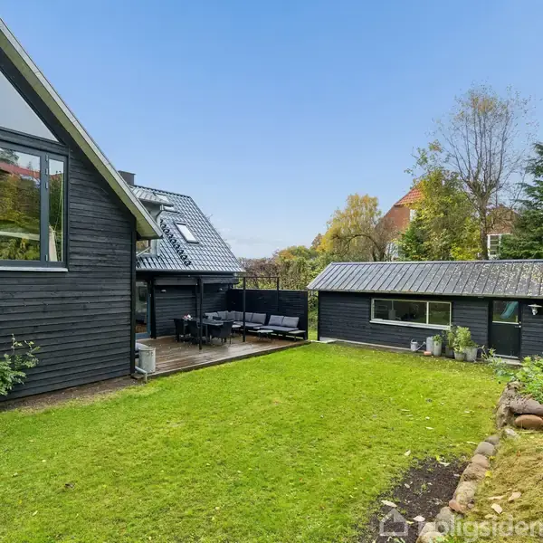 Sort træhus står stille ved siden af en velplejet græsplæne. Terrasse med stole og bord skaber en hyggelig atmosfære i haven, omgivet af træer og buske.