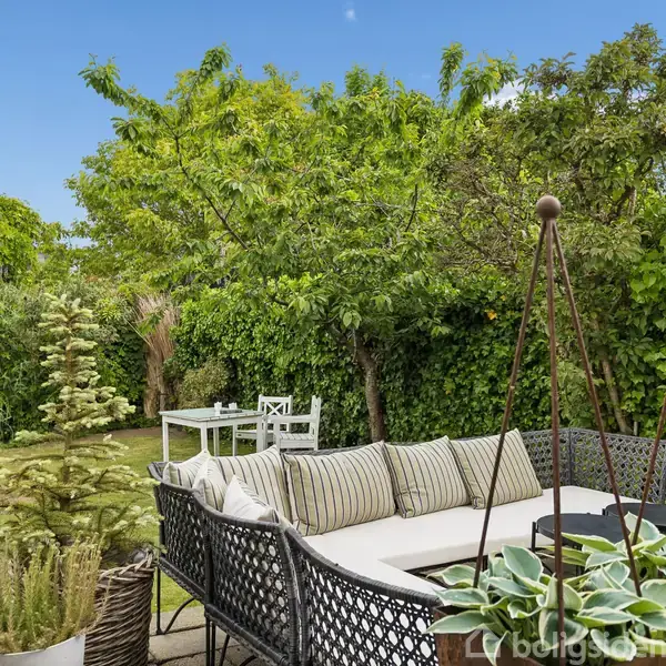 En udendørs sofa med puder står på en terrasse. I baggrunden ses en frodig have med grønne træer og en hvid havebænk under en blå himmel.