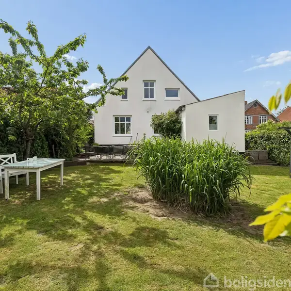 Hvidt hus står stille i en grøn have med græs og buske. Et hvidt havebord og stole er placeret på plænen. Solrigt vejr og blå himmel omkranser scenen.