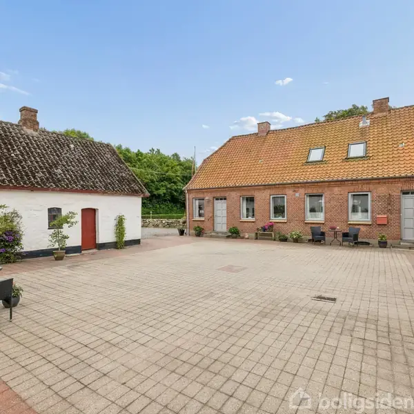 Stort gårdmiljø med fliser, omgivet af to røde murstensbygninger med skråtag. Havemøbler og planter på terrassen skaber en hjemlig atmosfære. Skyfri himmel og grønne træer i baggrunden.