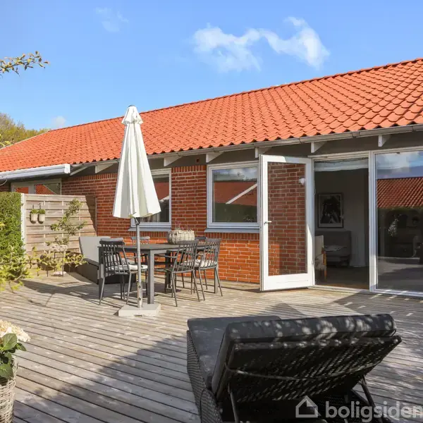 Et trædæk med en sort møbelgruppe og en parasol står foran et rødt, murstenshus med røde tegltage og store vinduer, omgivet af grønne planter og blå himmel.