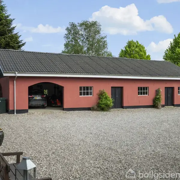 En rød bygning med tre garageporte og en indkørsel af grus. En bil er delvist synlig i den åbne garage. Omgivet af grønne træer og buske.