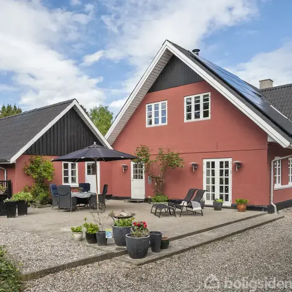 Rødt hus med hvide vinduer står stille i en solrig gårdsplads med plantekrukker og havemøbler. Baggrunden er en blå himmel og grønne træer i en landlig omgivelser.