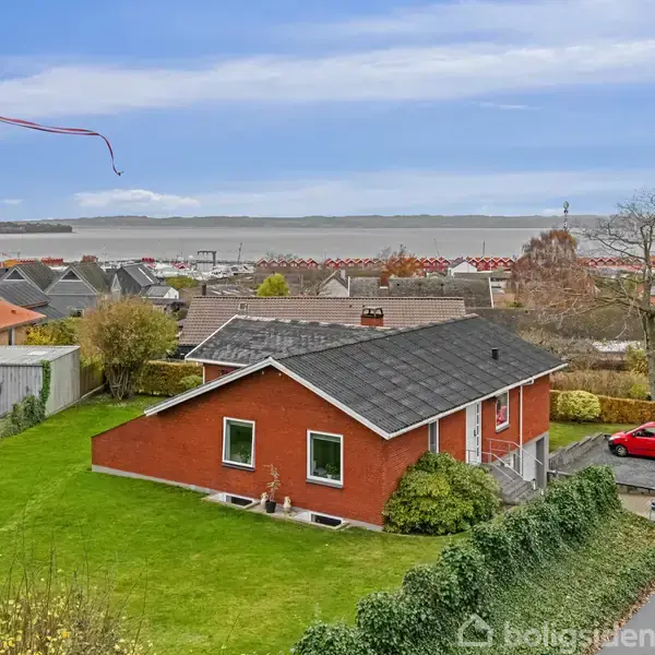 Et rødt hus står stille på en grøn græsplæne med udsigt over et vandløb og et kvarter. En rød bil er parkeret på vejen ved siden af huset.