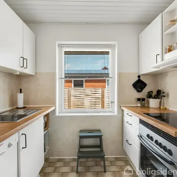 Et funktionelt køkken med hvide skabe og træbordplader. En ovn og kogeplade til højre, vask til venstre, og en lille skammel foran et vindue med udsigt til en træhegn.