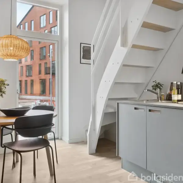 Rundt spisebord med sorte stole står under en hængende lampe ved siden af et køkken. En trappe fører opad. Store vinduer viser en rød bygning udenfor.