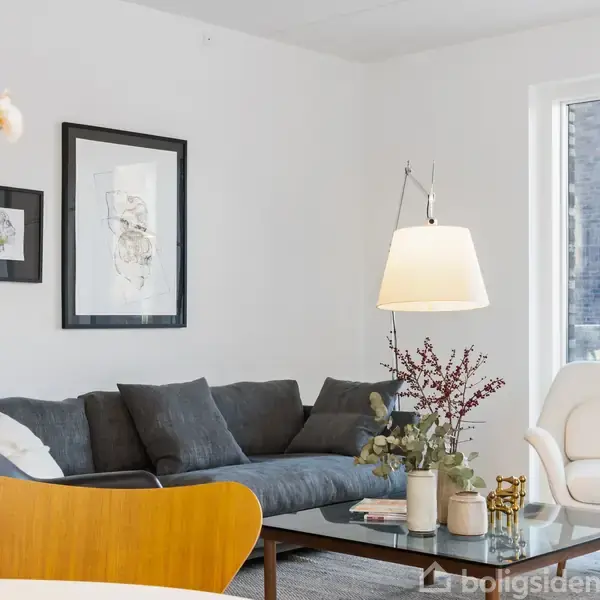 Grå sofa med puder står under vægudsmykning, flankeret af bord med planter og lamper, ved siden af hvid lænestol. Store vinduer giver udsigt til en bygning udenfor.
