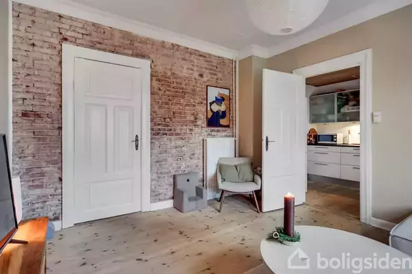 Dørleder værelset til køkkenet, mens en stol står ved en murstensvæg med kunst. Trægulv skaber hygge. En tændt lysestage står på bordet.