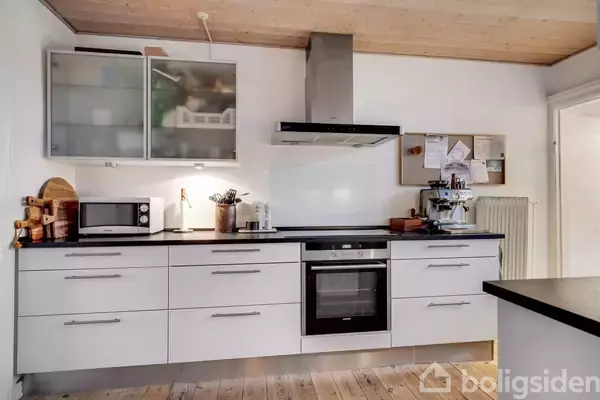 Et moderne køkken med hvide skabe og skuffer indeholder en indbygget ovn og emhætte. Til venstre står en mikrobølgeovn og køkkenredskaber. Baggrunden har et opslagstavle og træloft.