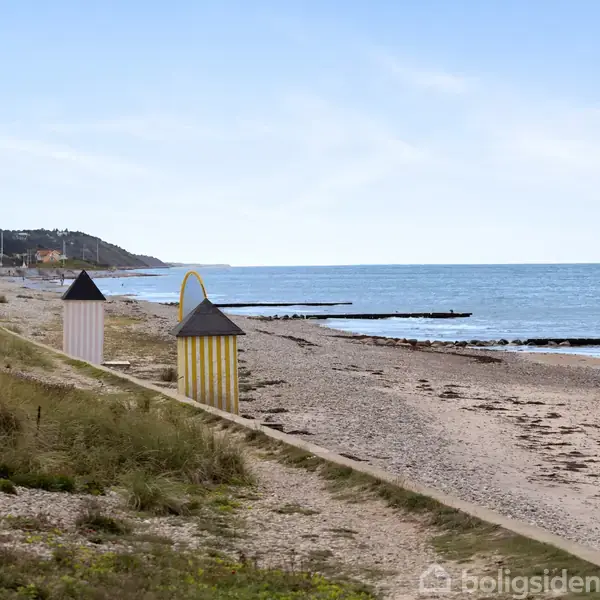 Strandhuse står stille på en åben sandstrand med små klitter og havet i baggrunden, mens en rolig kystlinje strækker sig under en lys, klar himmel.