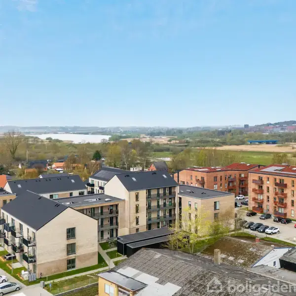 Flere moderne boligbygninger med flade tage står i et boligområde. Der er parkerede biler og grønne områder omkring bygningerne. I baggrunden ses en åben, landlig udsigt med fladt terræn.