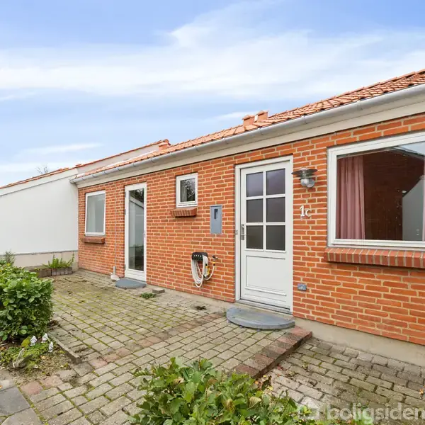 Rødt murstenshus med to vinduer og en hvid dør flankeret af en lille flisebelagt terrasse. En hæk og fliser løber langs huset, og himlen er blå med skyer.