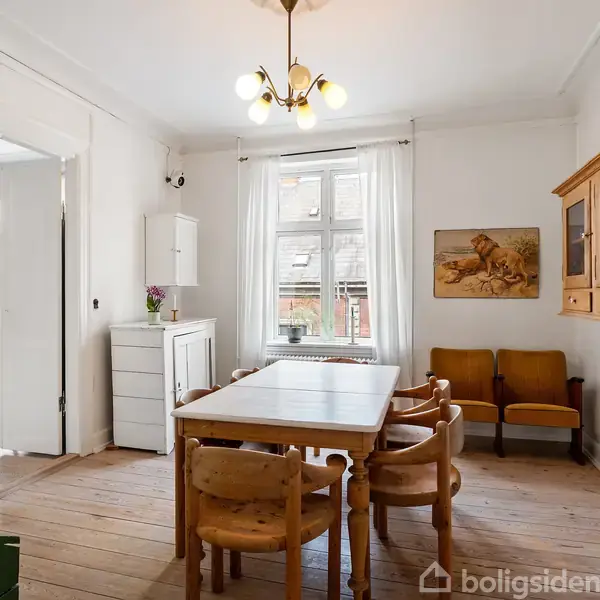 Spisebord med stole i et lyst rum. Reol hænger på væggen, maleri af to løver ved siden af. Vindue med hvide gardiner, døråbning fører til et andet rum.